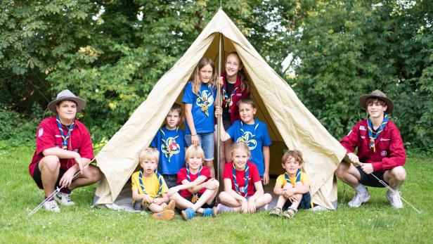 Die Pfadfinderinnen und Pfadfinder von Laa an der Thaya sind gemeinsam unter einem Zelt versammelt, das in der Natur steht. Zwei ältere Pfadfinder haben sich Außen am Zelt platziert.