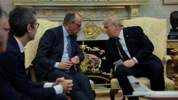 Donald Trump gestikuliert während eines Gesprächs mit einem Mann im Anzug in einem dekorierten Raum.