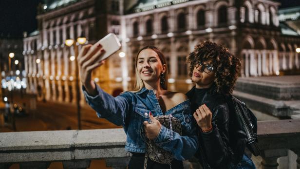 Zwei junge Frauen machen ein Selfie vor einem beleuchteten Gebäude in der Nacht.