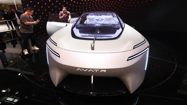 Futuristisches, weißes Konzeptauto mit offenem Dach und leuchtenden Linien, umgeben von Menschen und rotem Punktmuster im Hintergrund.