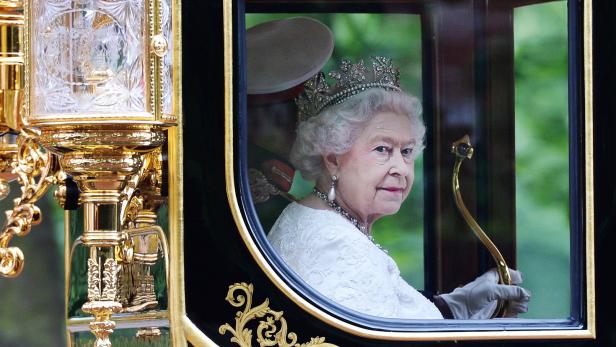 Königin Elisabeth II. mit Krone in einer Kutsche, neben ihr ein Gardist mit Hut.