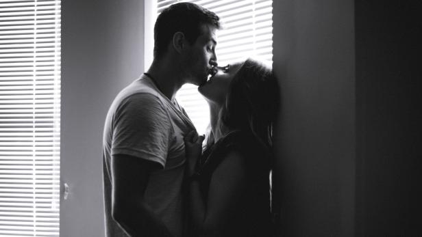 Ein Paar küsst sich leidenschaftlich vor einem Fenster mit Jalousien.