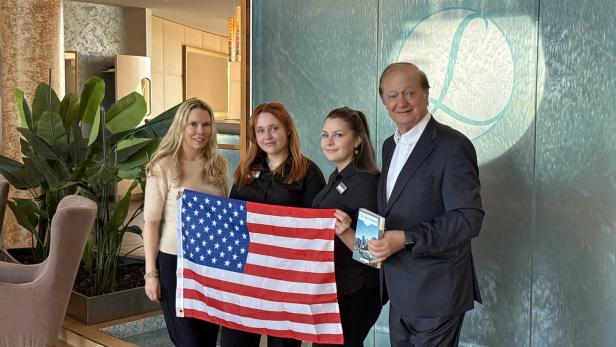 Vier Personen stehen nebeneinander, zwei davon halten eine US-Flagge, im Hintergrund eine Glaswand mit Hotel-Logo und Pflanzen.