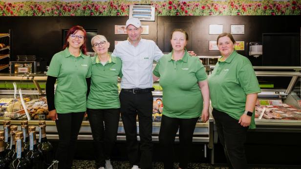 Fünf Personen in Arbeitskleidung stehen lächelnd vor einer Fleischtheke in einem Supermarkt nebeneinander.