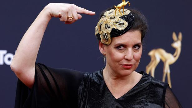 Frau mit schwarzem Kleid zeigt auf einen auffälligen Hut, der mit goldener Hirschfigur und Glitzer verziert ist.