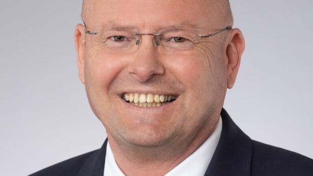 Ein lächelnder Mann mit Brille und Anzug vor grauem Hintergrund.