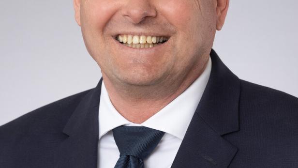 Ein lächelnder Mann mit Brille und Anzug vor grauem Hintergrund.