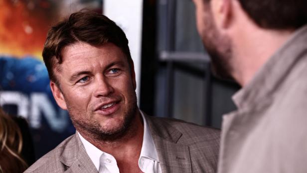 Luke Hemsworth mit Sakko spricht mit einer anderen Person.