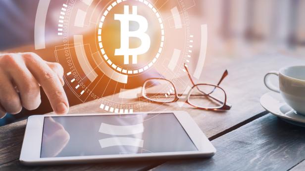 Eine Person tippt auf ein Tablet mit Bitcoin-Symbol, neben einer Tasse Kaffee und einer Brille.