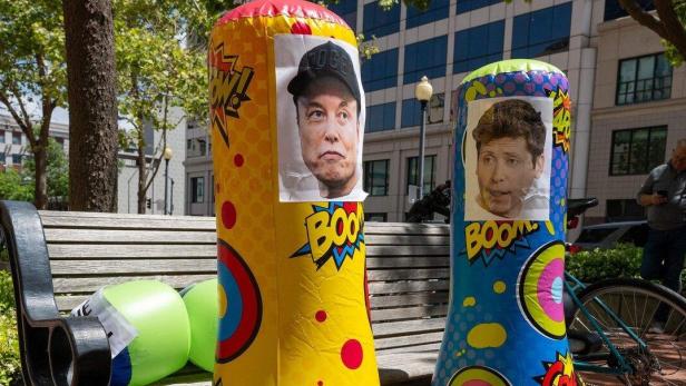 Auf einer Parkbank stehen zwei bunte Boxsäcke mit den Gesichtern von Elon Musk und einem weiteren Mann.