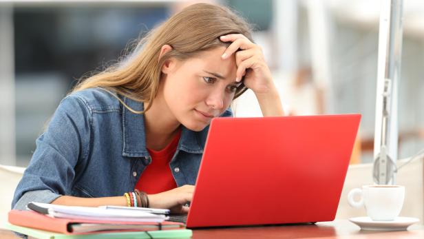 Eine junge Frau sitzt nachdenklich vor einem roten Laptop, umgeben von Unterlagen und einer Tasse Kaffee.