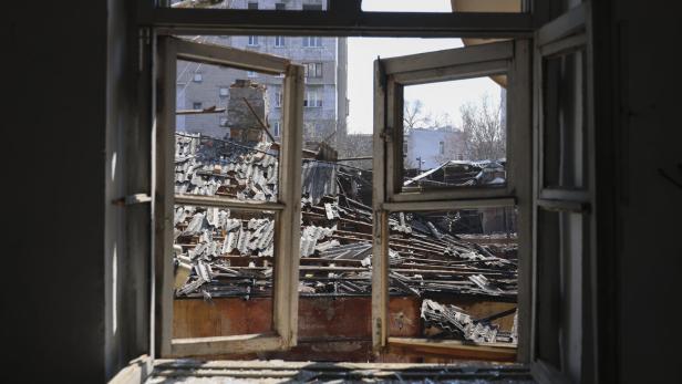 Durch ein geöffnetes Fenster sieht man ein zerstörtes Dach und umliegende Gebäude.