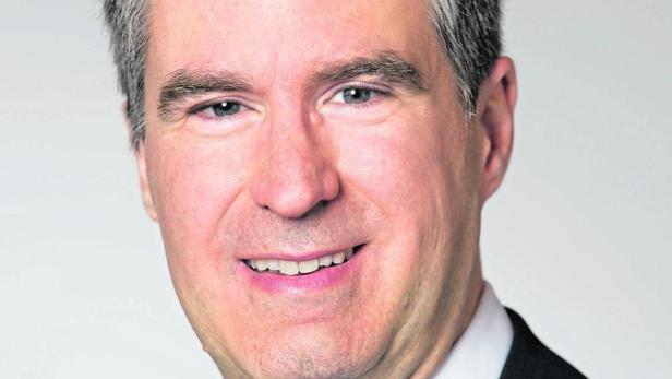 Ein Mann mit graumeliertem Haar lächelt in einem dunklen Anzug mit blau gestreifter Krawatte.