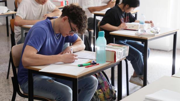 Mehrere Jugendliche sitzen an Einzeltischen in einem Klassenraum und schreiben konzentriert an Prüfungsunterlagen.