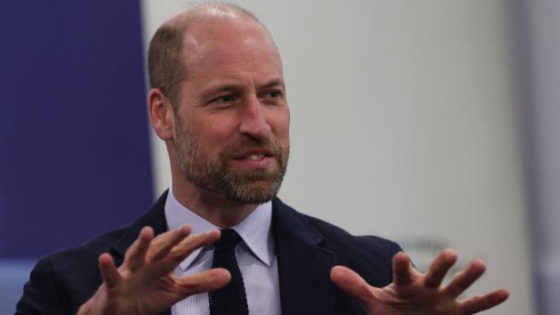 Prinz William gestikuliert in Anzug und Krawatte vor blau-weissem Hintergrund.