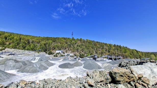 Steinbruch mit mehreren Kieshügeln, Förderbändern und angrenzendem Wald unter klarem, blauem Himmel.