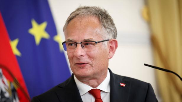 Ein Mann mit Brille und roter Krawatte spricht vor Mikrofonen, im Hintergrund sind EU- und Österreich-Flagge zu sehen.