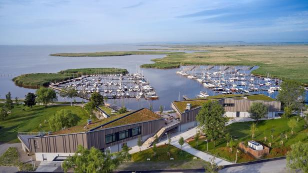 Zwei moderne Gebäude mit begrünten Dächern stehen an einem Yachthafen mit vielen Segelbooten, umgeben von Wasser und Schilf.