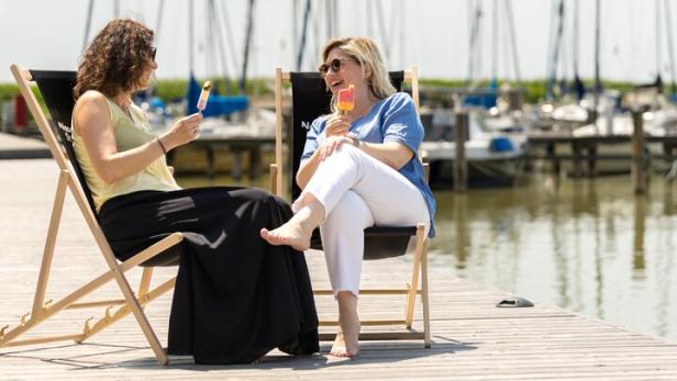 Zwei Frauen sitzen entspannt auf Liegestühlen am Steg, essen Eis und unterhalten sich, im Hintergrund Segelboote am Wasser.