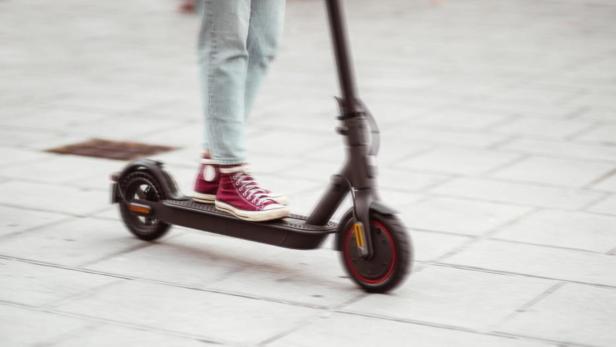 Person mit hellblauer Jeans und roten Sneakers fährt auf einem schwarzen E-Scooter über einen gepflasterten Weg.