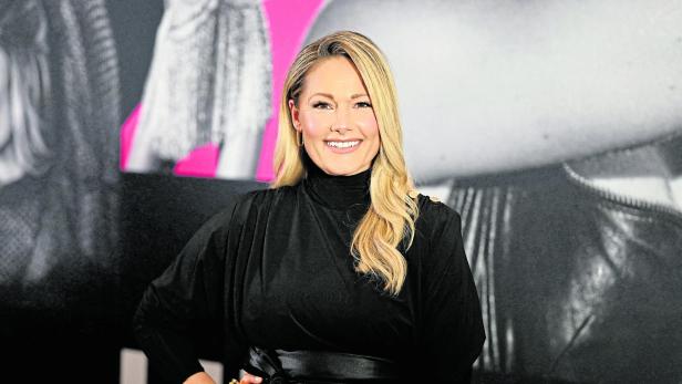 Helene Fischer lächelt in einem schwarzen Kleid mit Gürtel vor einem Hintergrund.