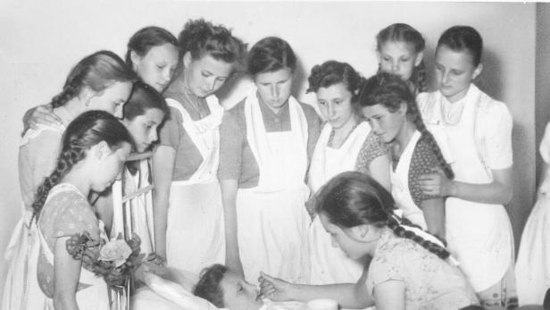 Mehrere Mädchen in Schürzen stehen um ein Bett, in dem ein weiteres Mädchen liegt und gefüttert wird.