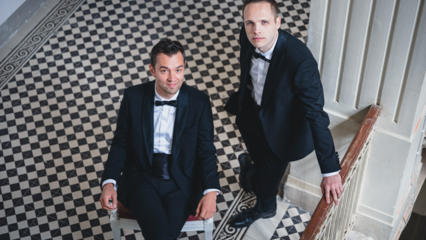 Nils Strunk und Lukas Schrenk im eleganten Anzug auf einer historischen Treppe, verantwortlich für die Neuinterpretation von „Die Fledermaus“ bei den Festspielen Reichenau 2026.