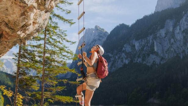 Eine Frau mit Rucksack klettert eine Leiter an einer Felswand über einem See hinauf.