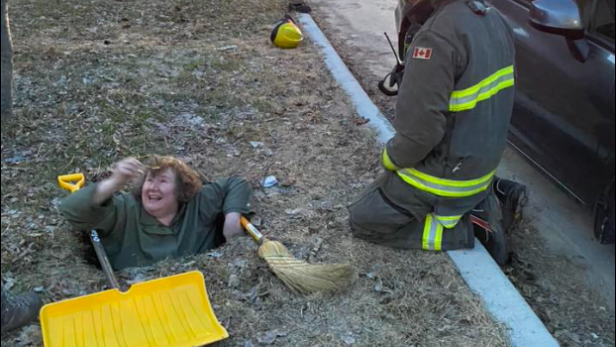 Frau steckt in einem Loch fest, Feuerwehrmann kniet daneben, Schneeschaufel und Besen liegen bereit.