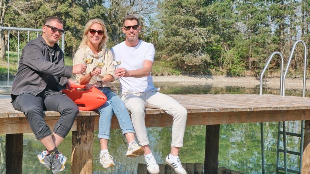 Drei Personen sitzen lachend auf einem Steg am Wasser, stoßen mit Gläsern an und tragen Sonnenbrillen.