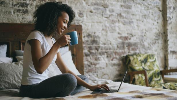 Frau sitzt auf ihrem Bett mit Kaffee und Laptop
