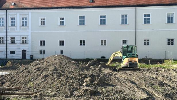 Ein Bagger steht auf einer Baustelle vor einem großen, weißen Gebäude mit rotem Dach.