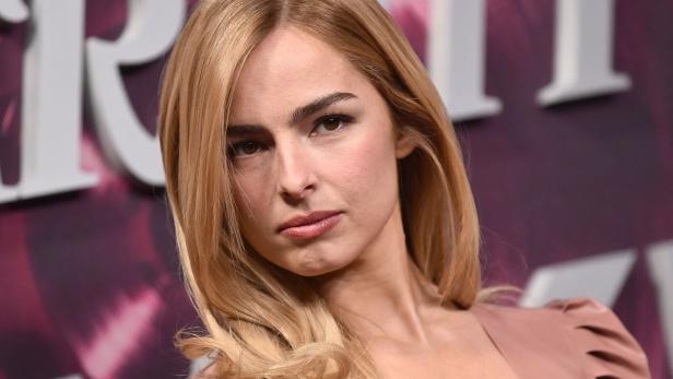 Eine Frau mit langen blonden Haaren und ernstem Blick steht vor einem lila-weißen Hintergrund.