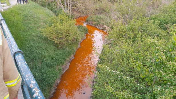 Oranger Fluss schlängelt sich durch grüne Vegetation, mehrere Personen stehen am Ufer.