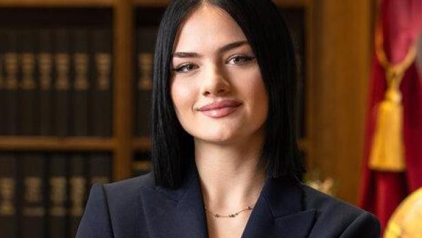 Junge Frau mit dunklen Haaren im Anzug steht vor einem Bücherregal mit juristischen Büchern und rotem Vorhang.