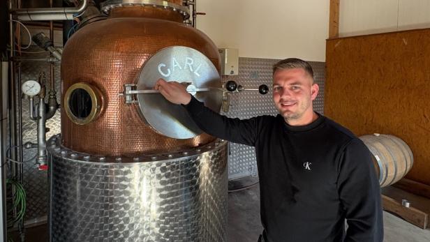 Ein Mann steht neben einer kupfernen Destille und hält den Griff einer runden Tür mit der Aufschrift „CARL“.