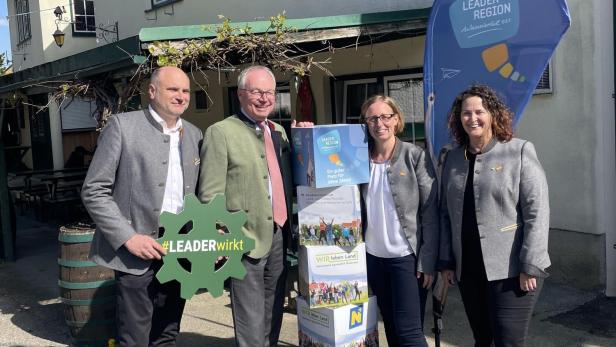 Vier Personen stehen vor einem Gasthaus neben einem LEADER-Region-Aufsteller und halten ein grünes Zahnrad mit der Aufschrift #LEADERwirkt.