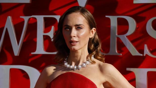 Emily Blunt mit Perlenkette und rotem, trägerlosem Kleid posiert vor rotem Hintergrund.
