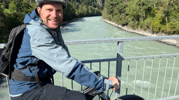 Ein Mann mit Helm und Rucksack fährt auf einem Fahrrad über eine Brücke mit Fluss und Wald im Hintergrund.
