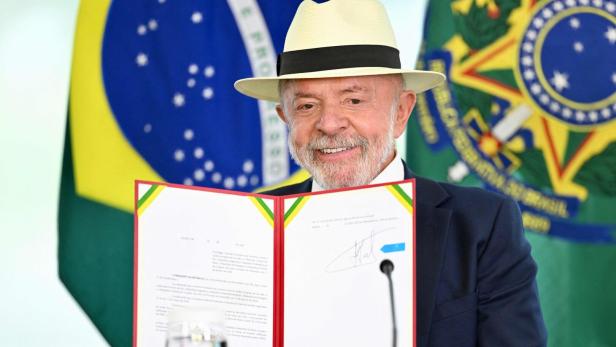 Ein älterer Mann mit Hut hält ein unterzeichnetes Dokument vor sich, im Hintergrund sind brasilianische Flaggen zu sehen.