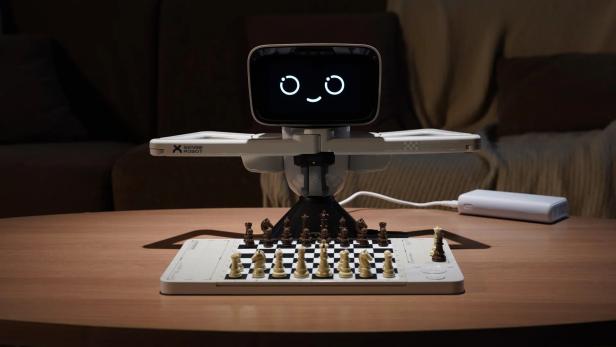 Ein Schachroboter mit Bildschirmkopf und Greifarmen steht vor einem Schachbrett auf einem Tisch.