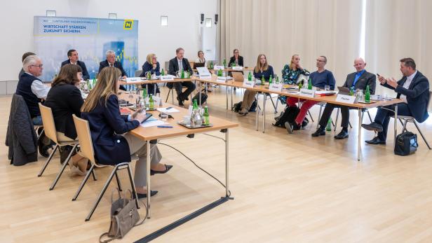 Viele Menschen sitzen in einem hell erleuchteten Raum vor einem Aufsteher des Wirtschaftslandes NÖ um einen Tisch.