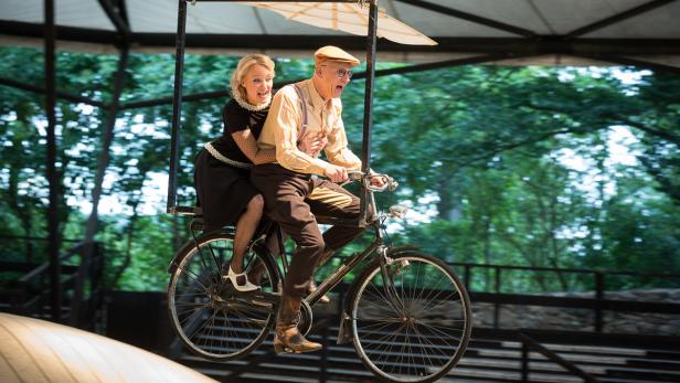 Ein Mann und eine Frau fahren gemeinsam auf einem dekorierten Fahrrad vor einer grünen Kulisse.