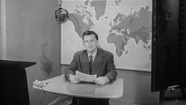 Ein Mann im Studio, mit Weltkarte im Hintergrund, hält ein Blatt Papier in den Händen.