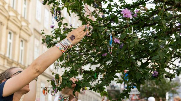 Eine Frau mit Armbändern hängt ein weiteres an einen Baum mit lila Blüten.