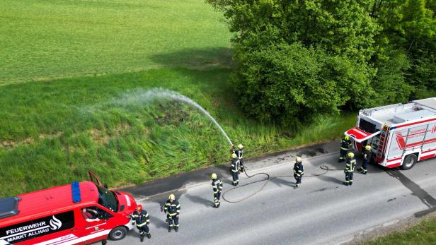 Mehrere Feuerwehrleute löschen mit einem Schlauch einen kleinen Brand am Straßenrand neben zwei Einsatzfahrzeugen.