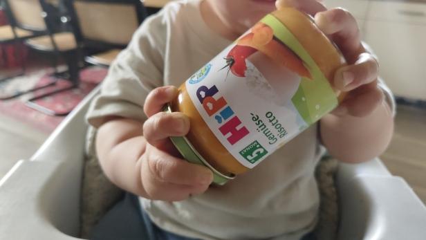 Ein Kleinkind hält ein Glas mit Hipp Gemüse Risotto Babynahrung in der Hand.