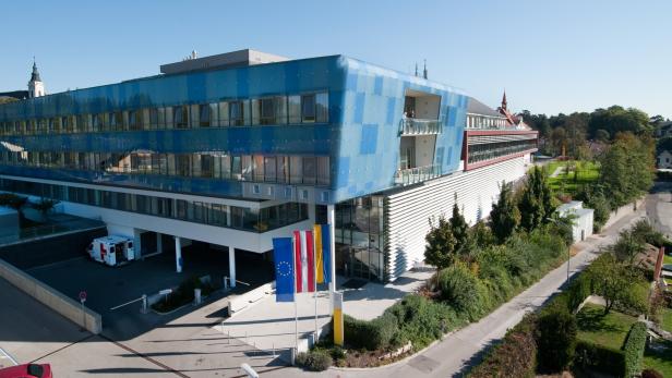 Modernes Gebäude mit blauer Glasfassade, davor drei Fahnenmasten und ein Rettungswagen, umgeben von Grünflächen und Straßen.