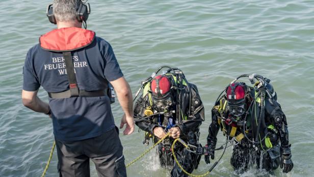 Zwei Feuerwehrtaucher steigen mit Ausrüstung aus dem Wasser, eine Person mit Headset steht am Ufer.
