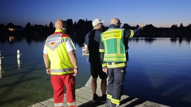 Drei Personen stehen auf einem Steg am Wasser, zwei davon in Einsatzkleidung, während die Dämmerung einsetzt.